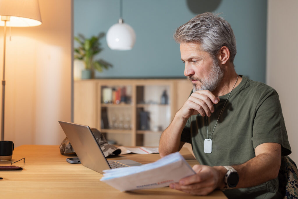 Gray-haired veteran in a green T-shirt and dog tags reviewing VA disability claim paperwork at his laptop at home.