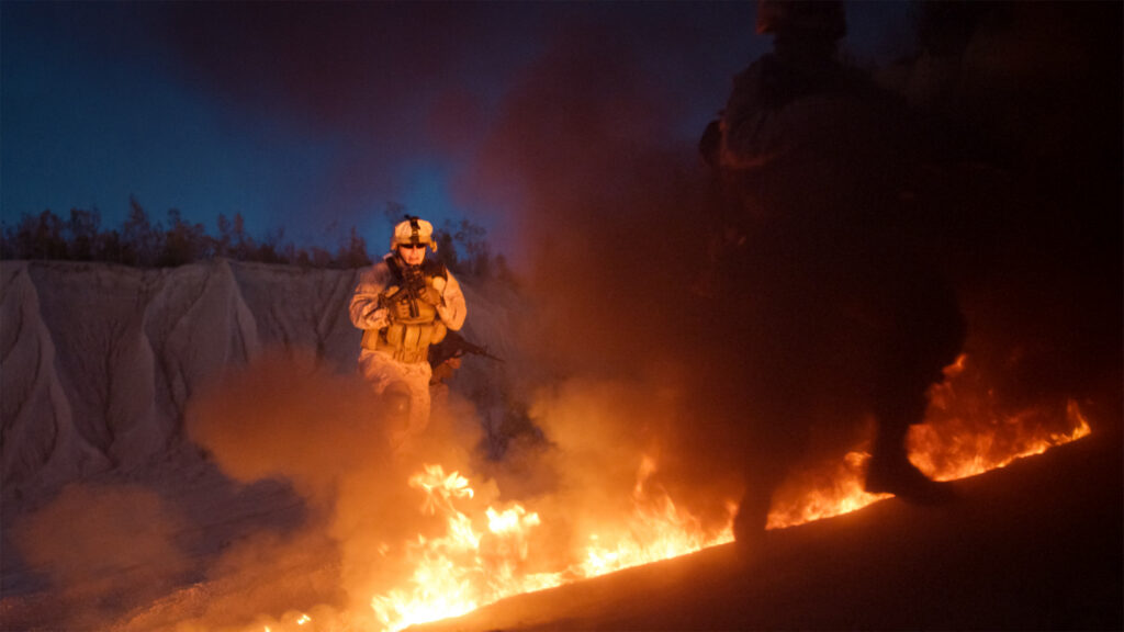 Service members advancing past open flames and smoke at night, illustrating burn pit and toxic exposure covered by the PACT Act