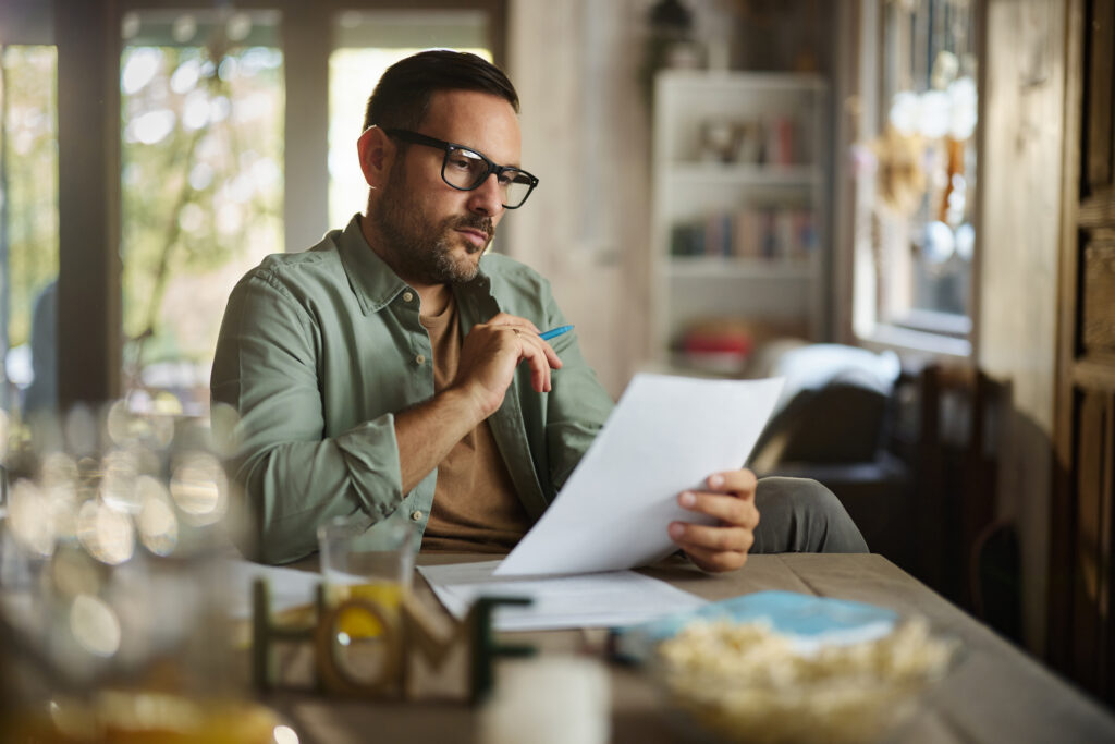 A man wearing glasses sits at a home dining table, closely reading a printed document with a pen in hand.