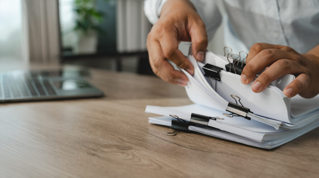 Hands sorting through binder-clipped stacks of paperwork next to an open laptop on a desk